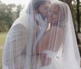Bride and groom smile and embrace under a sheer veil outdoors, surrounded by trees in soft daylight at a Louisiana wedding.