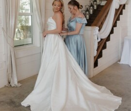 Bride in white gown stands by window as bridesmaid in blue helps; rustic room with stairs and candles, Louisiana wedding prep.