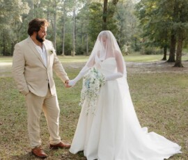 Bride in white dress and groom in beige suit hold hands outdoors at romantic Louisiana wedding surrounded by greenery.