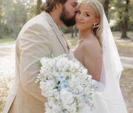 A groom in a beige suit kisses his smiling bride’s forehead outdoors, with trees and soft sunlight—romantic Louisiana wedding inspiration.