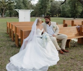 Bride in white gown and groom in tan suit smile on outdoor bench at their New Orleans wedding, surrounded by trees and benches.