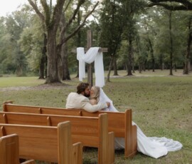 Bride and groom embrace on outdoor wooden pews under a cross with white fabric, surrounded by trees at a Louisiana wedding.
