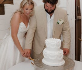 Smiling bride and groom cut a white two-tiered cake on a silver stand at their wedding reception.