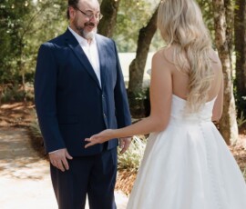 A groom in a navy suit smiles at his bride in a white dress on a sunny, tree-lined path at their Louisiana wedding.