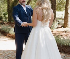 A groom in a navy suit holds hands with his bride in a white dress outdoors among trees and sunlight.