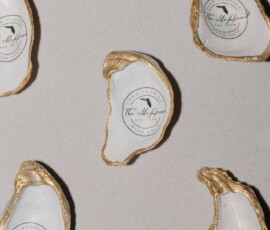 Gold-edged oyster shells with “The Albatross” labels, scattered on light fabric—unique New Orleans wedding place cards or favors.
