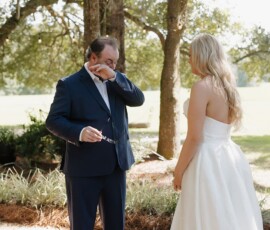 Emotional man in navy suit holding glasses faces bride in white dress during intimate outdoor wedding ceremony.