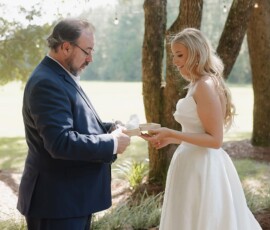 Bride in white gown and man in suit exchange cards outdoors under sunlit trees, perfect wedding inspiration for Louisiana.