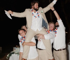 Men in tan suits and floral leis lift a laughing man at an outdoor nighttime Louisiana wedding celebration.