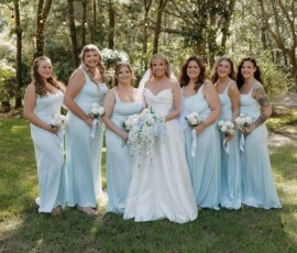 Bride in white gown with six bridesmaids in light blue, holding white bouquets, smiling outdoors at a Louisiana wedding.
