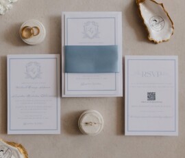 Elegant blue-accented wedding invitations, gold ring dishes, white florals, and QR RSVP card on a neutral surface.