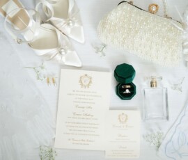 Wedding accessories flat lay with white heels, pearl clutch, gloves, invitations, ring box, perfume, and white florals.