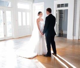 Bride in white gown and groom in black suit holding hands in a sunlit Louisiana wedding with wooden floors and large windows.