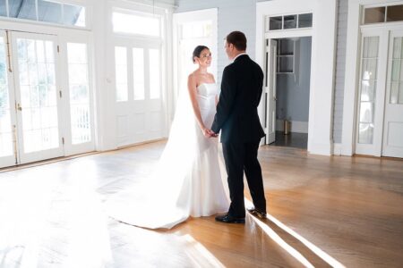 Bride in white gown and groom in black suit holding hands in a sunlit Louisiana wedding with wooden floors and large windows.