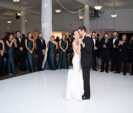 Bride and groom share a first dance on a white floor, surrounded by smiling bridal party in teal dresses and black tuxedos.