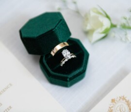 Gold wedding band and diamond engagement ring in green velvet box, with white flower and invitation in the background.