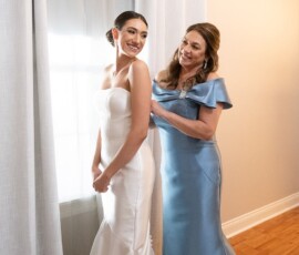 Bride in white dress smiles as woman in blue fastens her dress by a window—an intimate Gulf South wedding moment.