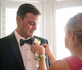 Woman in pink dress pins boutonniere on man in black tux at elegant New Orleans wedding by bright windows.