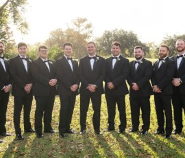 Nine men in black tuxedos and bow ties smile outdoors on grass, sunlight filtering through trees at a Louisiana wedding.