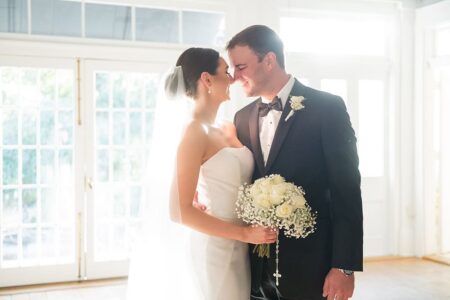 Bride in white dress with bouquet smiles beside groom in tuxedo, celebrating their joyful New Orleans wedding indoors.