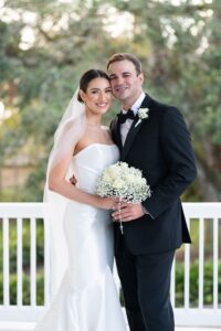Bride in white gown and veil with groom in black tux, smiling on porch with bouquet; classic Louisiana wedding scene.