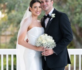 Bride in white gown and veil with groom in black tux, smiling on porch with bouquet; classic Louisiana wedding scene.