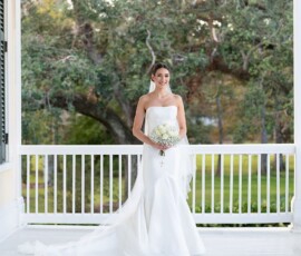 Bride in white gown on porch with bouquet, surrounded by greenery, reflecting timeless New Orleans wedding elegance.