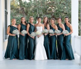 Bride in white and seven bridesmaids in dark green stand with bouquets on a porch, classic Louisiana wedding charm.