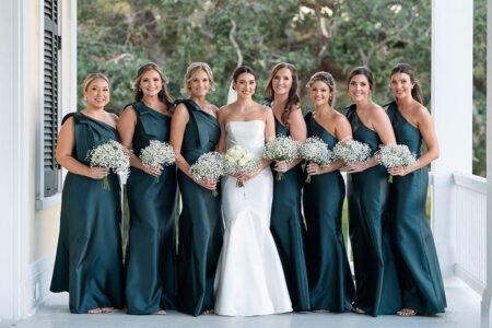 Bride in white gown with seven bridesmaids in dark green, all holding white bouquets, smiling on a porch with trees behind.
