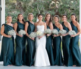 Bride in white gown with seven bridesmaids in dark green, all holding white bouquets, smiling on a porch with trees behind.