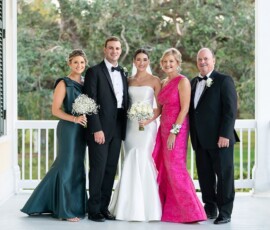 Five elegantly dressed people pose on a white porch at a New Orleans wedding, smiling with bouquets and greenery behind them.