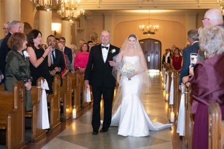 A bride in a white dress walks down a candle-lit church aisle with an older man, surrounded by guests at a classic New Orleans wedding.