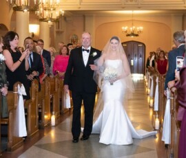 A bride in a white dress walks down a candle-lit church aisle with an older man, surrounded by guests at a classic New Orleans wedding.