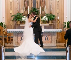 Bride and groom share a kiss at a New Orleans church wedding, surrounded by guests, flowers, and candles.