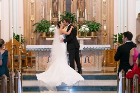 Bride and groom share a kiss at a New Orleans church wedding, surrounded by guests, flowers, and candles.
