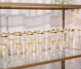 Champagne glasses with gold rims arranged on a glass shelf, with white drapery and soft lighting for a romantic wedding touch.