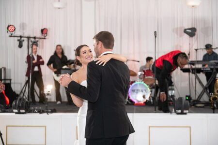 Bride and groom dance as a live band sets up on stage in a bright, elegant Louisiana wedding venue.