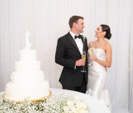 Bride and groom in formalwear smile, holding champagne; white wedding cake and flowers in front, drapery and lights behind.