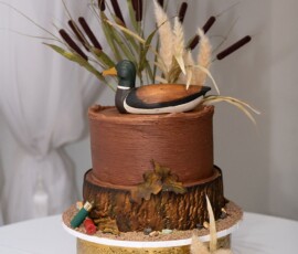 Chocolate wedding cake with duck topper, reeds, cattails, and shotgun shells on gold stand with pine and dried plants.