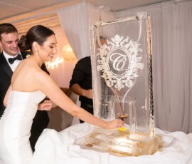 Bride pours drink from C-etched ice sculpture as two men in tuxedos assist at elegant Gulf South wedding reception.