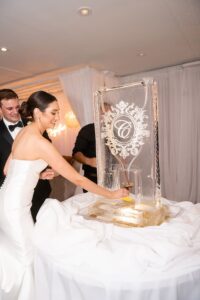 Bride pours drink from C-etched ice sculpture as two men in tuxedos assist at elegant Gulf South wedding reception.