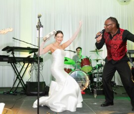 Bride in white gown dances with tambourine beside singer in red vest at lively Louisiana wedding with band and green stage lights.