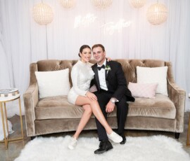 Smiling couple in formal attire on a beige sofa, with wedding decor and a “Better Together” neon sign in the background.
