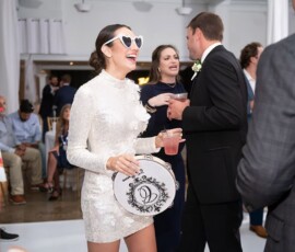 Woman in sparkly white dress and heart sunglasses smiles with tambourine at lively indoor wedding; guests mingle in background.