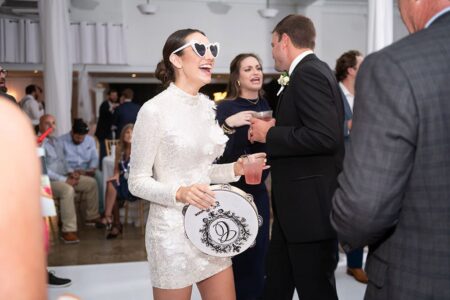 Woman in sparkly white dress and heart sunglasses smiles with tambourine at lively indoor wedding; guests mingle in background.