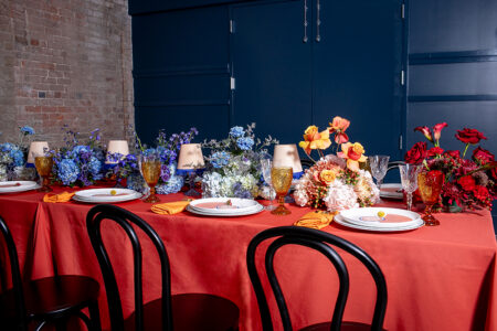 A red tablecloth covers a dining table with floral centerpieces, yellow napkins, crystal glasses, black chairs, and garnished plates.