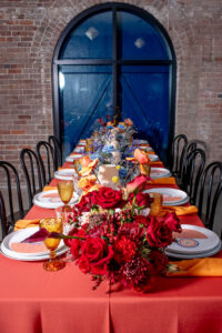 Long table with orange tablecloth, red and yellow flowers, gold goblets, black chairs, brick wall, and arched window.