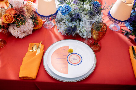 Colorful table with orange tablecloth, gold utensils, floral centerpieces, crystal glasses, and vintage lamp lighting.