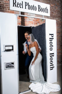 Bride in white gown and veil and groom in blue suit smile with fun glasses in a brick-walled photo booth.