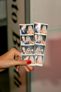 Hand with red nails holding photo booth strips of a man and woman making funny faces and posing with props by a curtain backdrop.